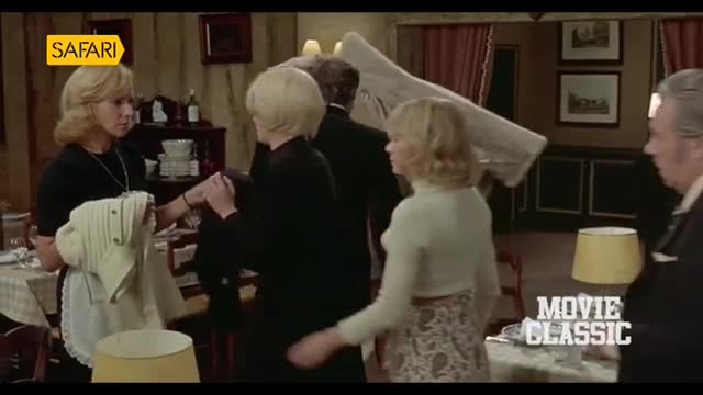 A woman in a black dress holds a white apron, seemingly attending to another woman's arm. Behind them, a man carries a large, folded white object through a dining room.
