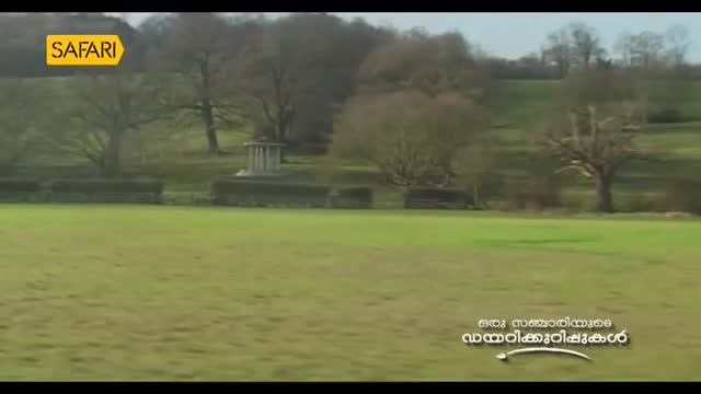 A wide, grassy field stretches out, with bare trees and a small stone structure on a distant hillside. The Safari TV logo sits in the corner, and Malayalam text appears at the bottom of the screen.
