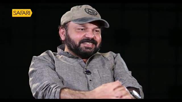The man, wearing a cap and a grey shirt, is speaking, his face lit by the studio lights. A Safari TV logo is visible in the corner, suggesting this is a program filmed in India.
