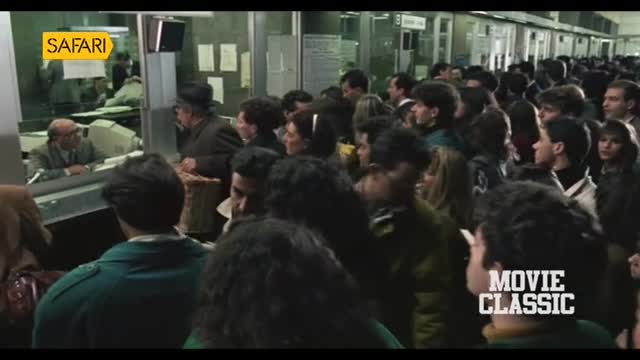 A dense crowd presses towards a row of ticket windows, the line stretching far back. People are clustered together, their faces turned towards the clerks behind the glass.
