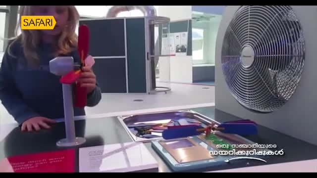 A woman holds a small model wind turbine in front of a large fan, presumably for a segment on Safari TV. The blades of the model are red and spin slowly, while the fan behind it remains still.
