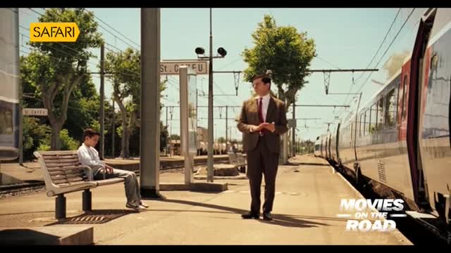 A man in a suit stands on the platform, while a train is pulled up beside him. A young man sits on a bench, seemingly waiting.
