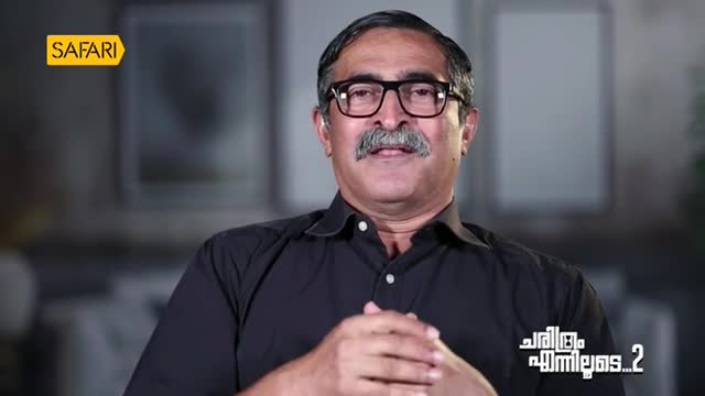 A man in a black shirt and glasses, speaking on a Safari TV show from India, is looking directly at the camera. His hands are clasped, and a logo for the show is visible in the corner.
