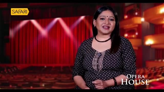 A woman in a patterned top stands in front of a red curtain, speaking to the camera for Safari TV's "Opera House" segment. The stage lights are focused on the curtain, and the seats of the theater are visible in the background.
