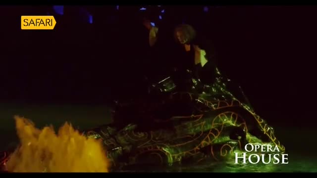 A performer, lit by a spotlight, stands on a glowing, ornate structure that resembles a dragon.  Yellow mist billows from the front as Safari TV presents the Opera House production.

