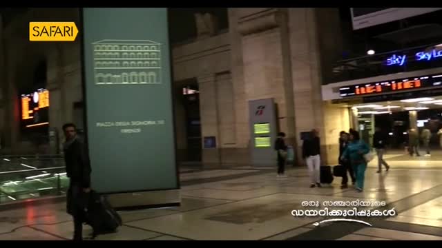A man with a suitcase walks past a large sign in a European train station, likely for a segment on Safari TV. Other people are scattered around, some pulling luggage, some just passing through.
