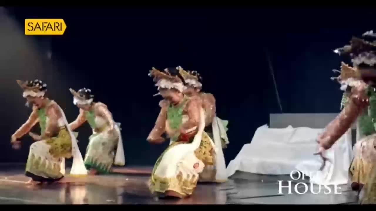 Dancers in elaborate headdresses and long, patterned skirts move across the stage, their arms gracefully extended. The performance, likely from India and broadcast by Safari TV, unfolds under a spotlight.
