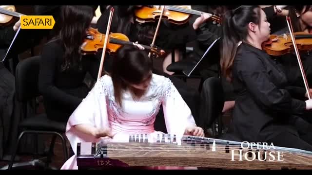 A woman in a flowing pink and white dress is intently plucking the strings of a large, wooden instrument, while a full orchestra accompanies her. The Safari TV logo is visible, and the scene appears to be broadcast from an opera house.
A woman in a flowing pink and white dress is intently plucking the strings of a large, wooden instrument, while a full orchestra accompanies her. The Safari TV logo is visible, and the scene appears to be broadcast from an opera house.