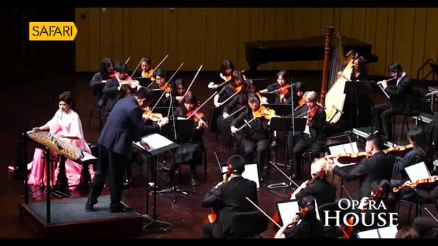 A woman in a pink gown plucks strings on a traditional instrument, while the conductor leads the orchestra with a violin. The Safari TV logo is visible as the performance unfolds at the Opera House.
A woman in a pink gown plucks strings on a traditional instrument, while the conductor leads the orchestra with a violin. The Safari TV logo is visible as the performance unfolds at the Opera House.