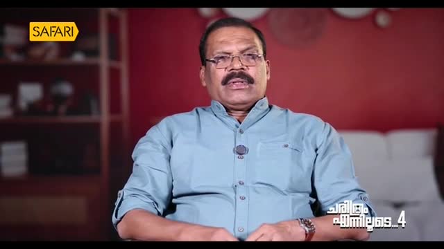 A man with a mustache and glasses is speaking directly to the camera, his mouth open as if mid-sentence. The Safari TV logo is visible in the corner, and the background is a blurred red wall.
