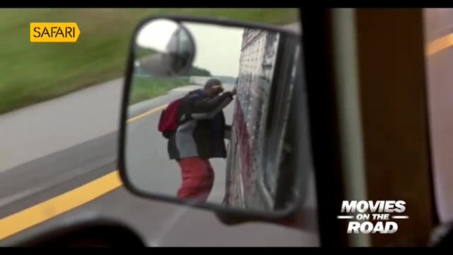 A man in red pants and a black jacket is clinging to the side of a truck, his back to the camera. The scene is reflected in the side mirror, a snippet from "Movies on the Road" on Safari TV.

