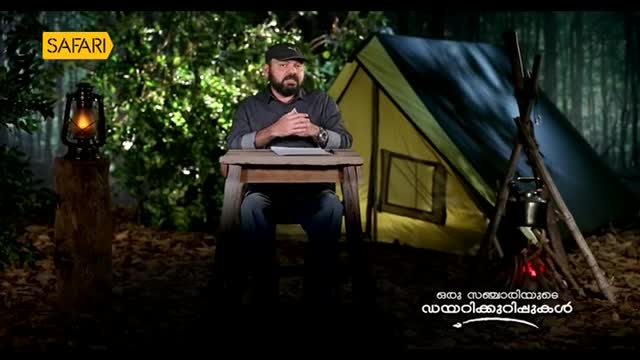 A man sits at a small table, a notepad in front of him, speaking to the camera. Behind him, a tent and a campfire glow in the darkness of the woods, as seen on Safari TV from India.
