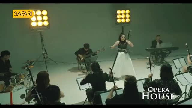 The woman in the white dress plays a stringed instrument, while others in the ensemble accompany her. The scene is lit by stage lights, and the "Safari" logo is visible.
