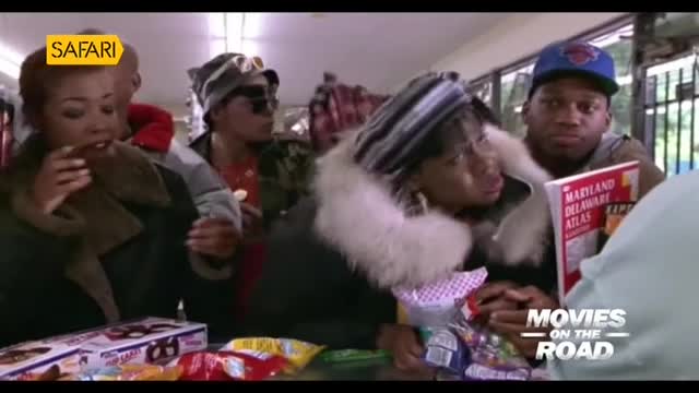 A group of people are gathered around a counter, some eating snacks. One man holds a map, while others seem to be looking at the contents of a bag.
