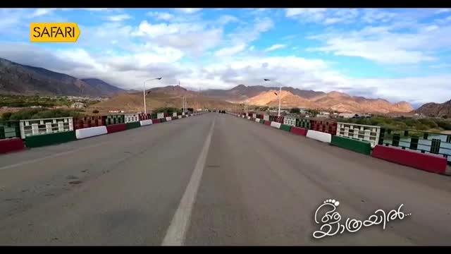 A line of cars moves steadily across a wide bridge, their headlights catching the afternoon sun. The Safari TV logo is visible in the upper left corner.