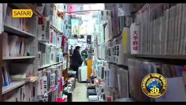 Two people navigate a narrow aisle packed with books and papers. A white suitcase sits on the floor, and a yellow object is propped against a shelf.