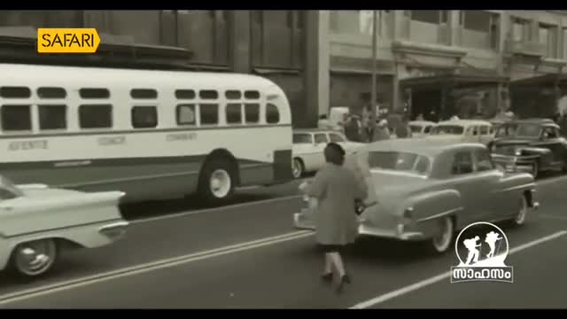 A white bus rolls past, its windows reflecting the city street. A woman in a coat hurries across the road, her heels clicking on the pavement.