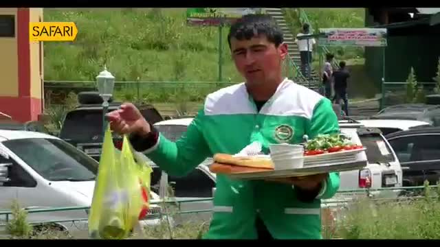 A man in a green and white uniform holds out a yellow plastic bag in his left hand. He balances a tray with a sandwich, salad, and a cup of sauce on his right.