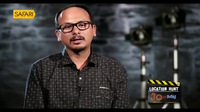 A man with glasses and a mustache speaks directly to the camera. Behind him, studio lights stand tall, and a clapperboard announces "Location Hunt."