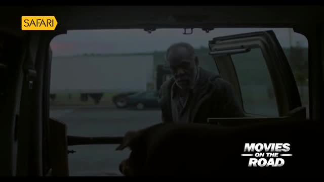 A man with a gray beard leans into the back of a vehicle, his face shadowed. A large, dark shape, possibly an animal, is partially visible in the foreground.