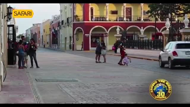 A white SUV turns down a street lined with brightly colored buildings. A child in a pink dress stumbles as a man in red shorts pulls her along.