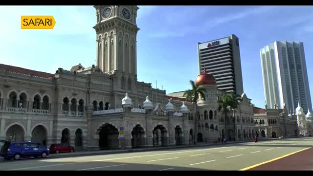 A blue van and a red car are parked on the side of a wide road. The grand, ornate facade of a historic building stretches out before me, its clock tower reaching towards the bright sky. A blue van and a red car are parked on the side of a wide road. The grand, ornate facade of a historic building stretches out before me, its clock tower reaching towards the bright sky.