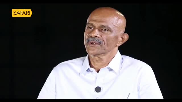 A man with a mustache and a shaved head speaks into a microphone, his white shirt crisp. The Safari TV logo appears in the top left corner. A man with a mustache and a shaved head speaks into a microphone, his white shirt crisp. The Safari TV logo appears in the top left corner.