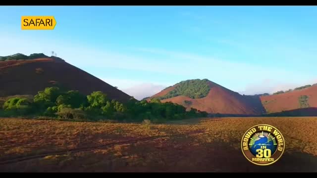 The sun rises over rolling hills in India, casting long shadows across a dry, grassy plain. A faint track winds through the landscape, hinting at journeys taken.