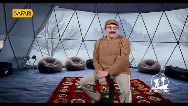 A man in a brown sweater and cap sits on a stool inside a geodesic dome, speaking to the camera. The backdrop behind him is a hazy, wintry landscape, hinting at a travelogue segment for Safari TV.