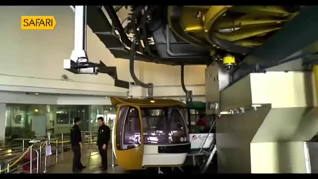 A yellow cable car idles at the station, its doors open. Two uniformed men stand nearby, observing its readiness for the Safari TV broadcast.