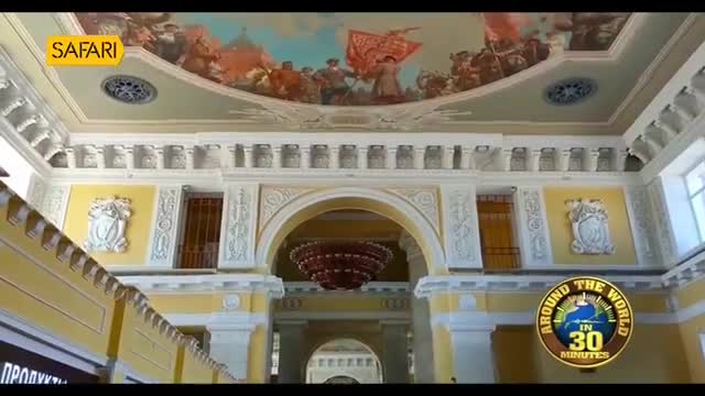 The grand hall of a train station, adorned with a vibrant mural of a procession, unfolds before me. A massive, multi-tiered chandelier hangs from the ornate ceiling, its deep red catching the light.