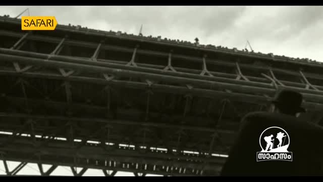 A man in a cap stands on a platform beneath a massive, dark structure. The Safari TV logo is visible on his shirt as he looks up.