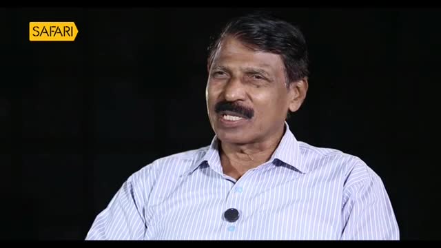 A man with a mustache speaks into a microphone, his light blue striped shirt visible. A yellow "Safari" logo sits in the upper left corner.