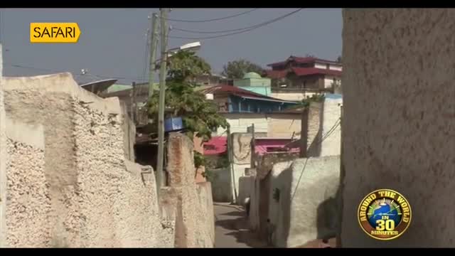 A narrow alleyway winds between rough, textured walls. Buildings with brightly colored roofs rise beyond, their facades a patchwork of faded paint and exposed brick.