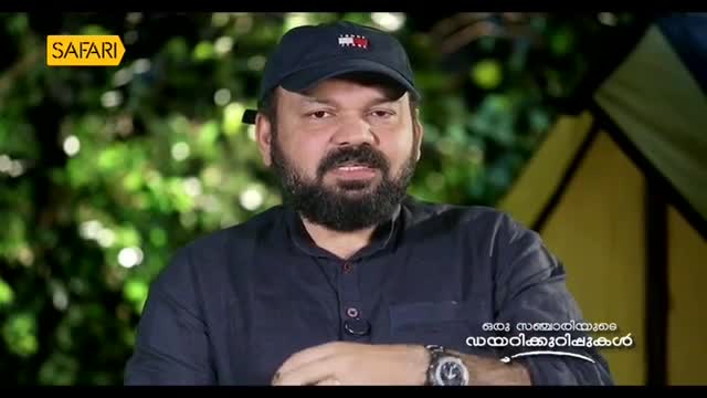A man with a beard and a cap speaks, his watch glinting under the lights. Behind him, lush green foliage and a yellow tent suggest an outdoor setting, perhaps for a Safari TV program.