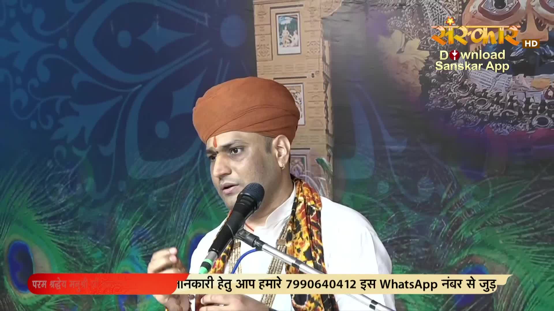 A man in a turban speaks into a microphone, his hand gesturing as he addresses the audience. Behind him, a peacock feather design decorates the background, and the Sanskar TV logo is visible.
