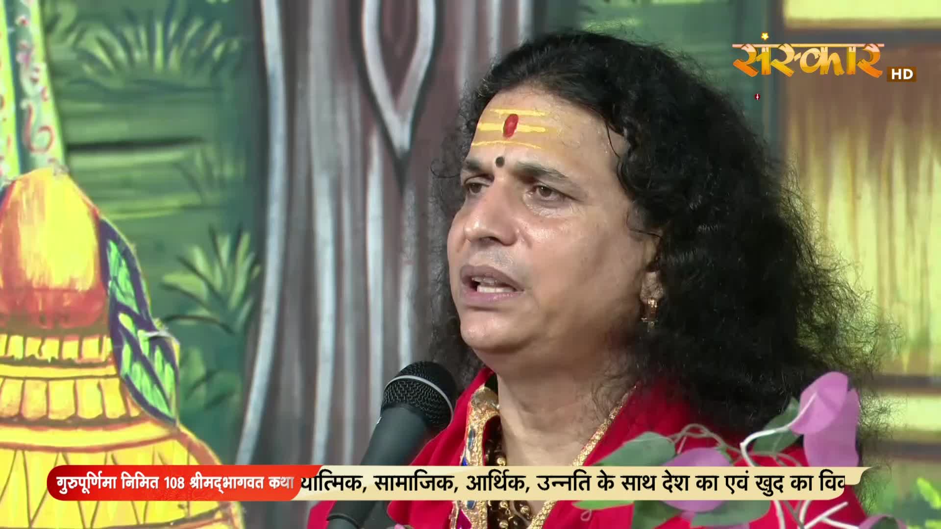 A man with a red tilak speaks into a microphone, his words broadcast on Sanskar TV in India. The backdrop is a colorful painted design, and text scrolls along the bottom of the screen.

