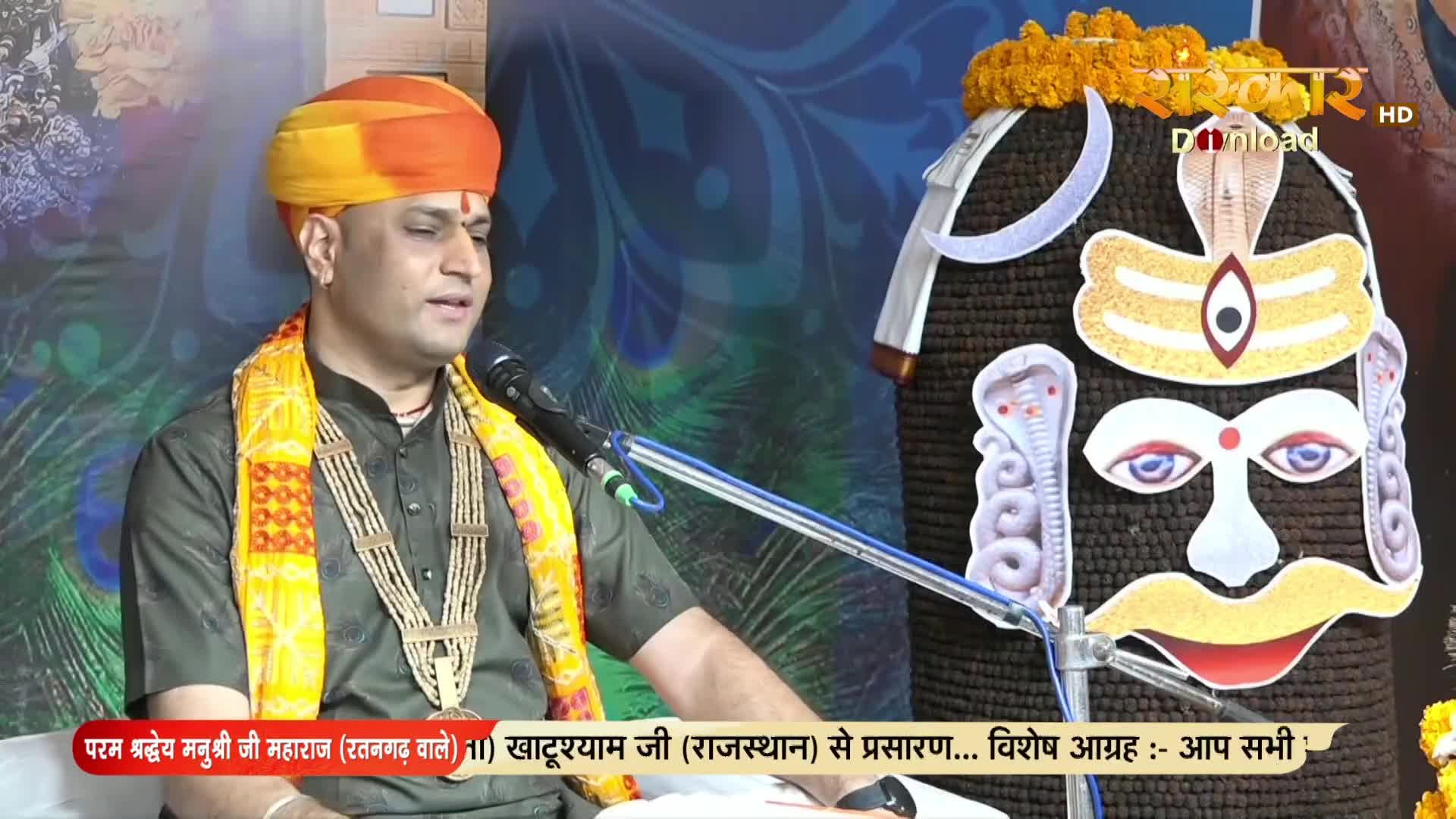 A man in a turban and beaded necklace speaks into a microphone, likely on Sanskar TV. Behind him, a large, stylized face of a deity is decorated with flowers.
