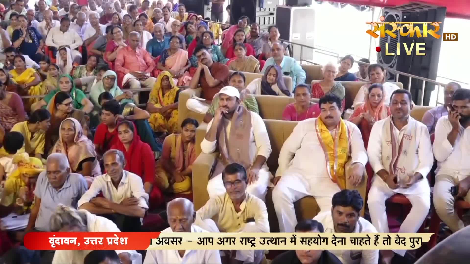 A large crowd sits in a hall, many in traditional Indian clothing, some looking towards the camera, others chatting quietly. The broadcast logo for Sanskar TV is visible, and the screen shows a live event in Vrindavan, Uttar Pradesh, India.

