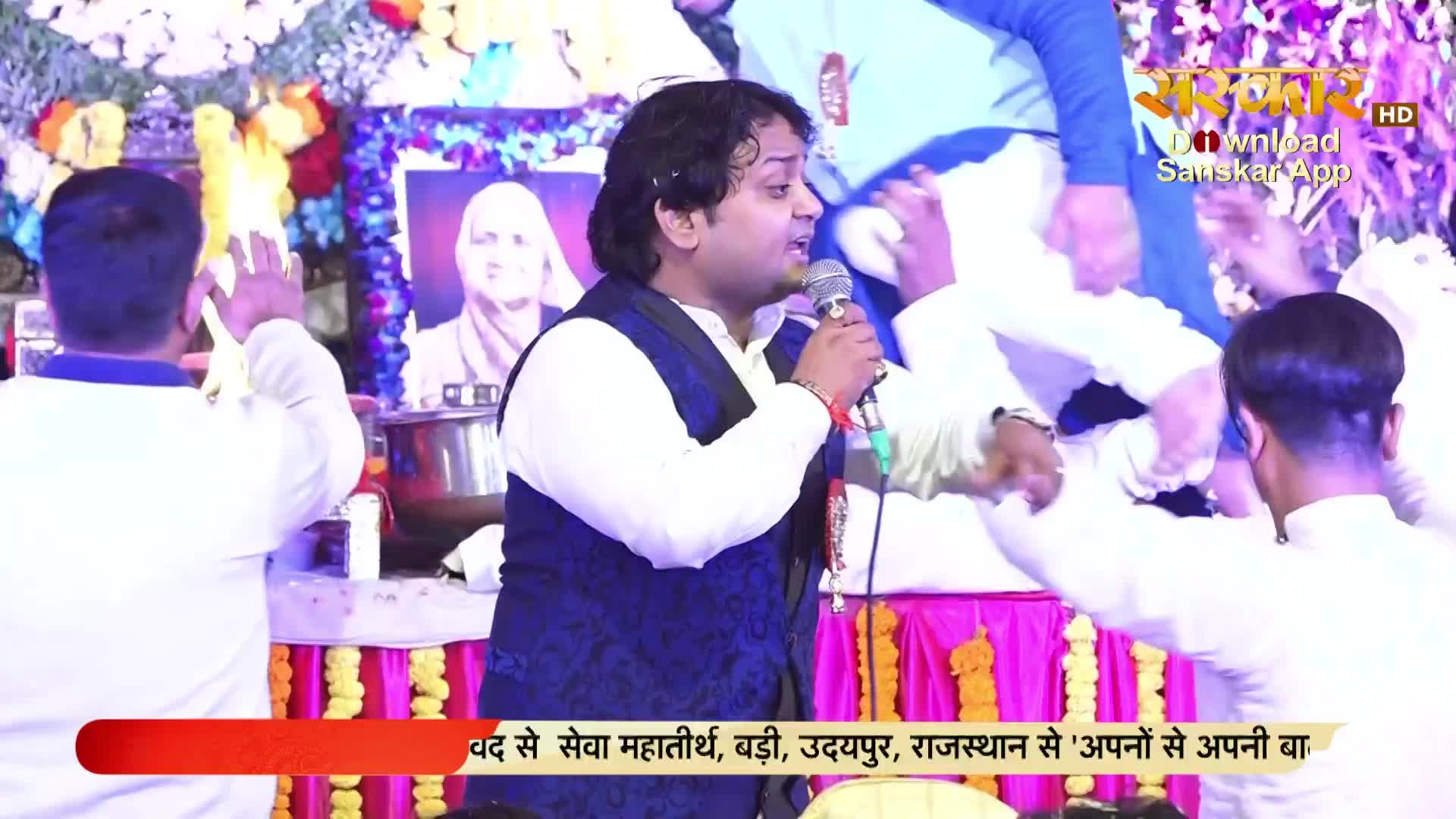 A man in a patterned vest sings into a microphone, gesturing with his right hand.  Around him, people in white shirts raise their hands, seemingly in a devotional setting, which is being broadcast by Sanskar TV.
