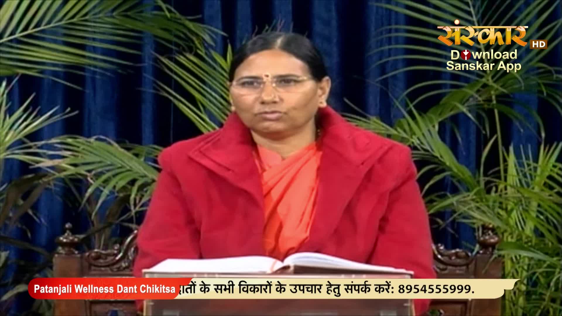 A woman in a red coat sits behind a desk, looking directly at the camera. The backdrop includes plants and a logo for Sanskar TV, a channel popular in India.
