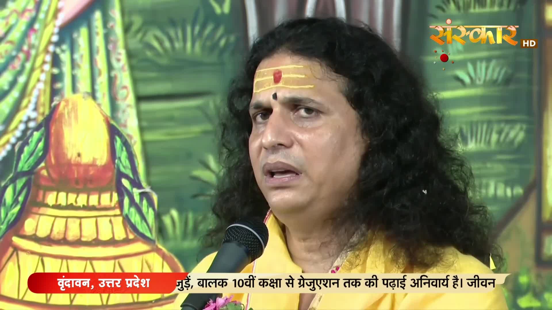 A man with long, dark hair and a mark on his forehead speaks into a microphone. The Sanskar TV logo and text from Vrindavan, Uttar Pradesh, are visible on the screen.
