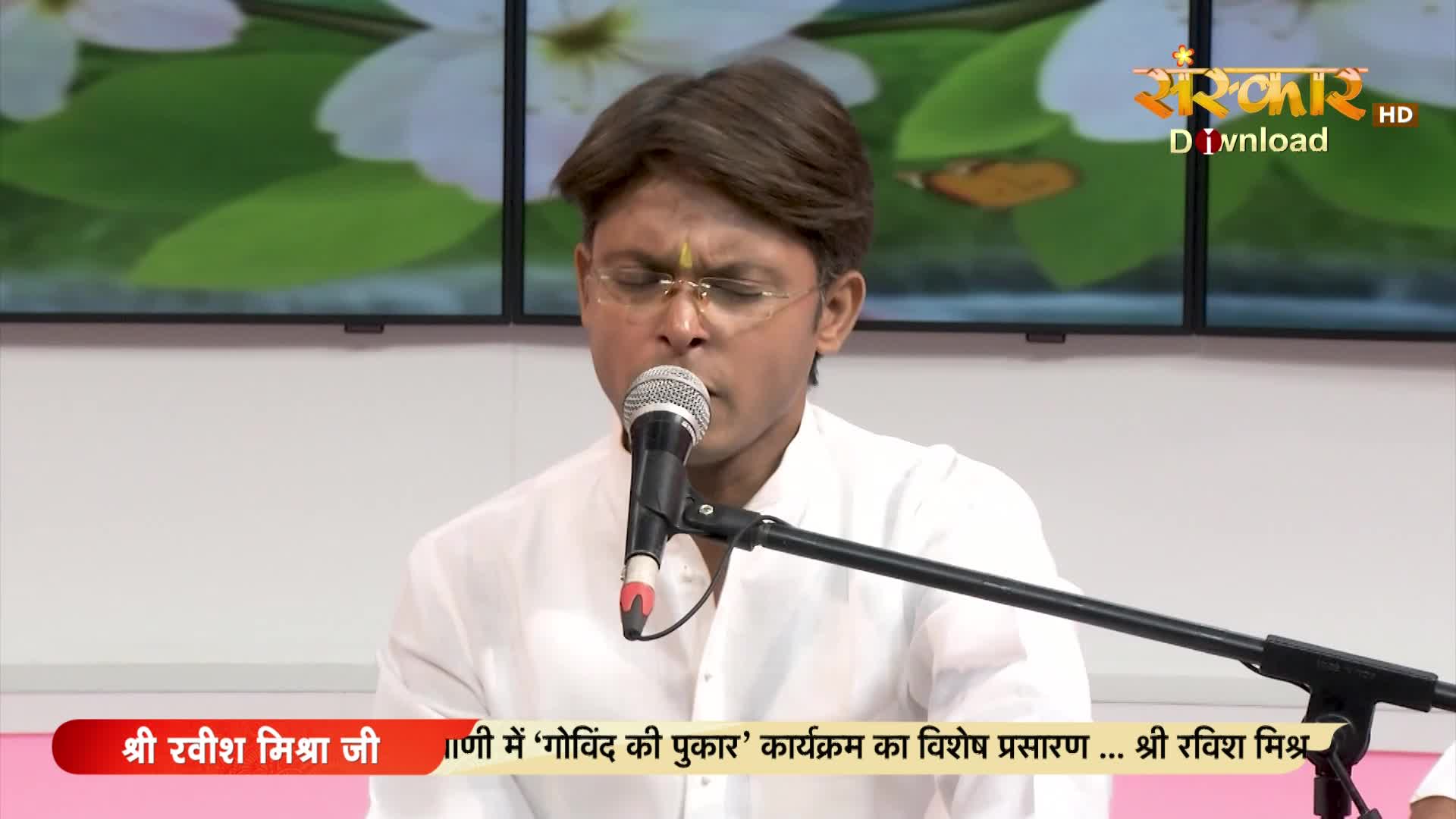 A man in a white shirt, wearing glasses, is singing into a microphone on a set for Sanskar TV. The bottom of the screen displays text in Hindi, identifying the program as "Govind Ki Pukar."
