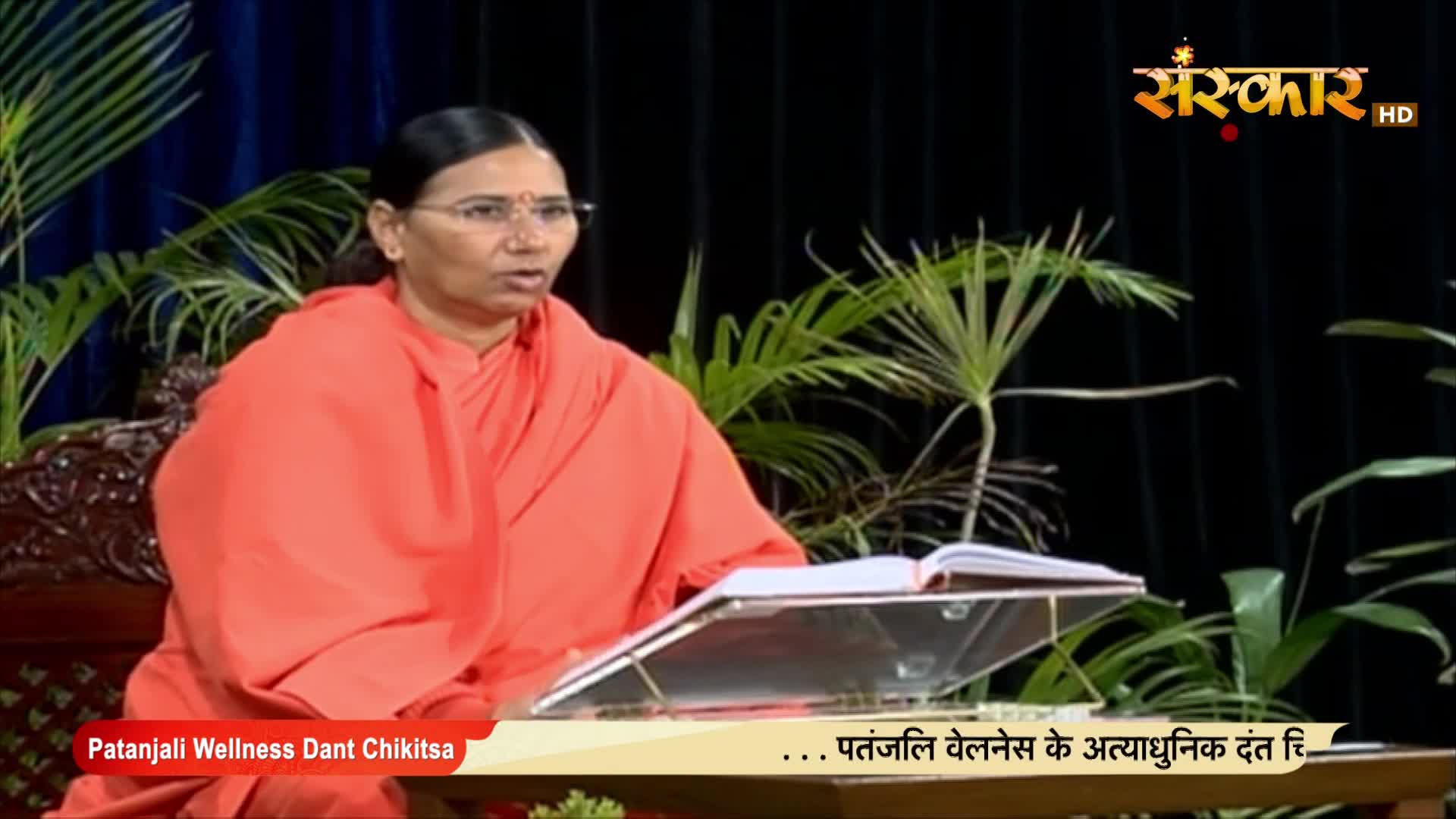 A woman in an orange robe is speaking, her words directed towards the camera, with a book open on a lectern in front of her. The Sanskar TV logo is visible, and the broadcast appears to be about wellness.

