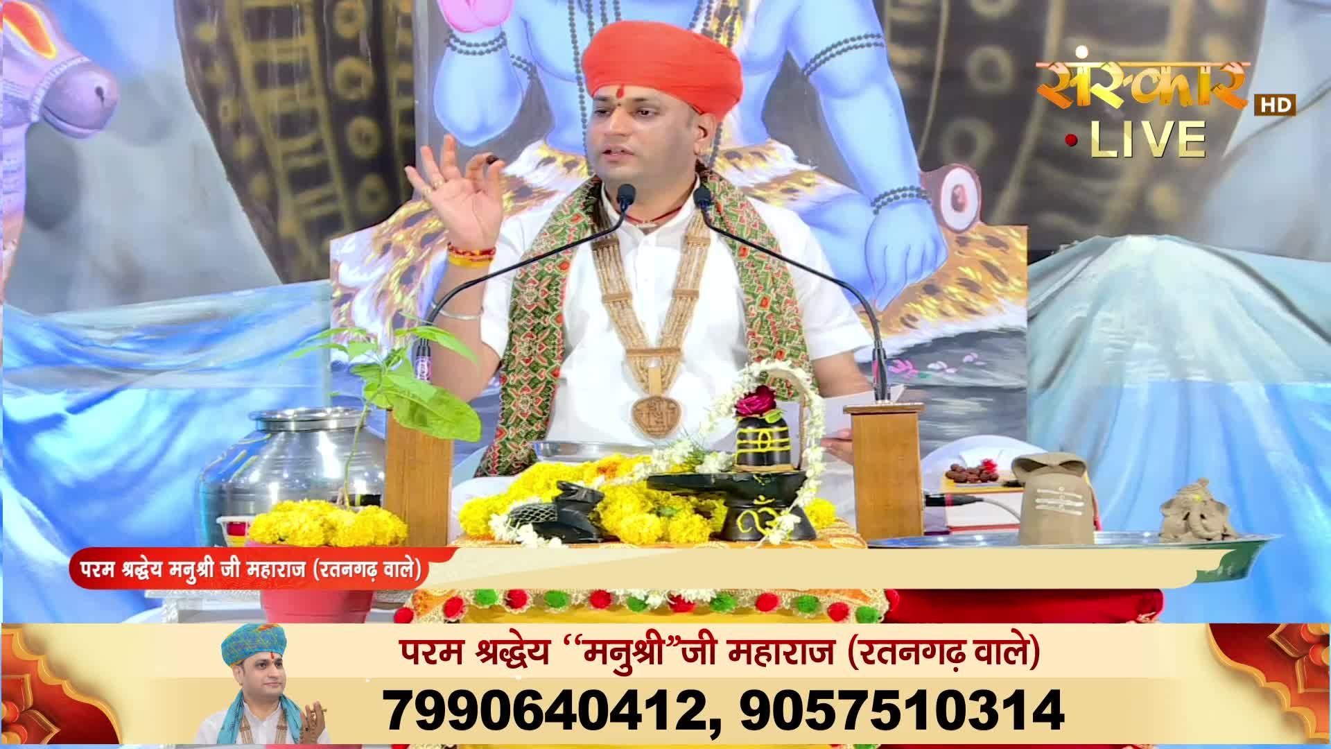 A man in a turban gestures with his hand while speaking into a microphone, the Sanskar TV logo visible in the corner. He's seated at a decorated altar, with a backdrop featuring a blue deity.
