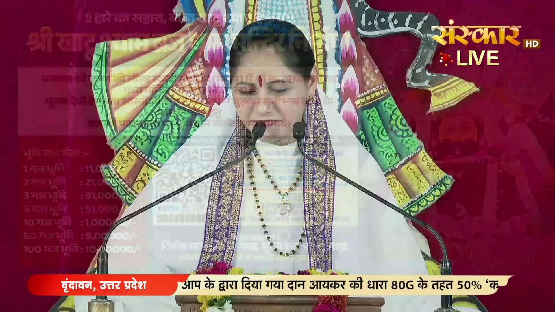 A woman in white speaks into two microphones, her face framed by a screen with Hindi text and a QR code. The Sanskar TV logo is visible in the upper right corner.
