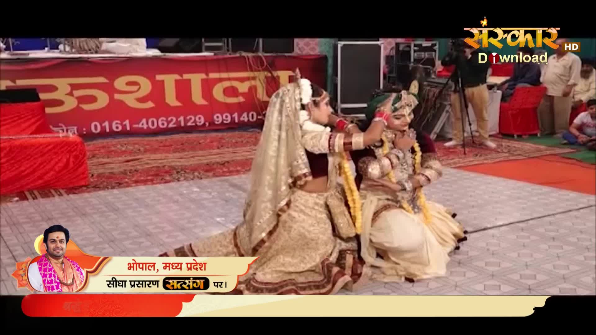 Two performers in ornate costumes are on a stage, with one adjusting the other's headpiece. The scene is being broadcast live on Sanskar TV, with a banner indicating the event is taking place in Bhopal, India.
