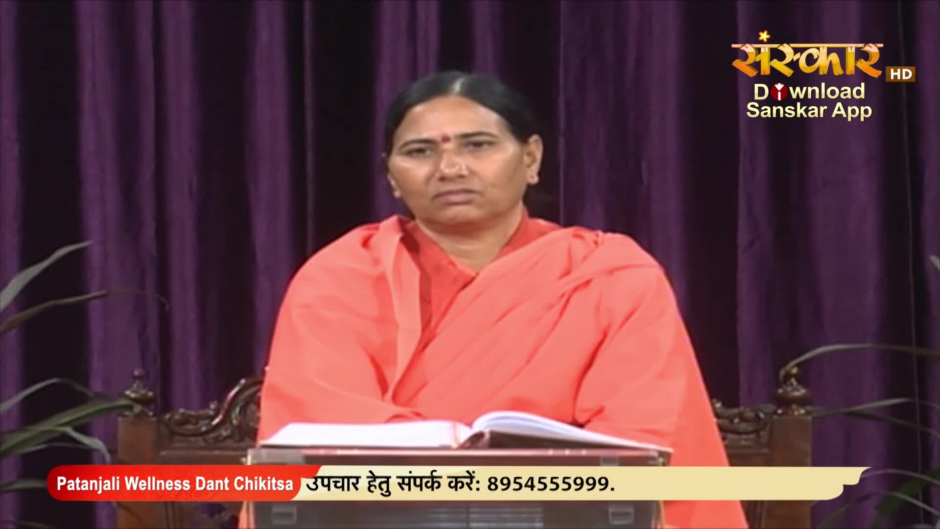 A woman in an orange robe sits at a table, looking directly at the camera. The Sanskar TV logo and a phone number for Patanjali Wellness are visible on the screen.
