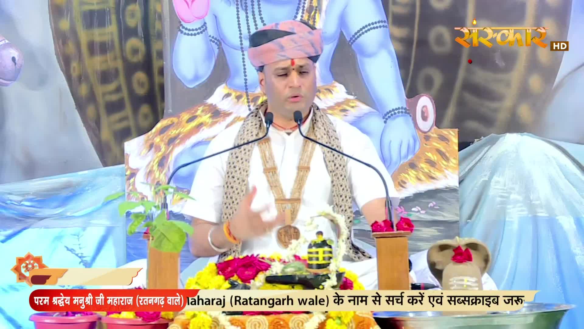 A man in a turban and white shirt gestures while speaking into two microphones. Behind him, a backdrop with a blue deity and the Sanskar TV logo is visible.
