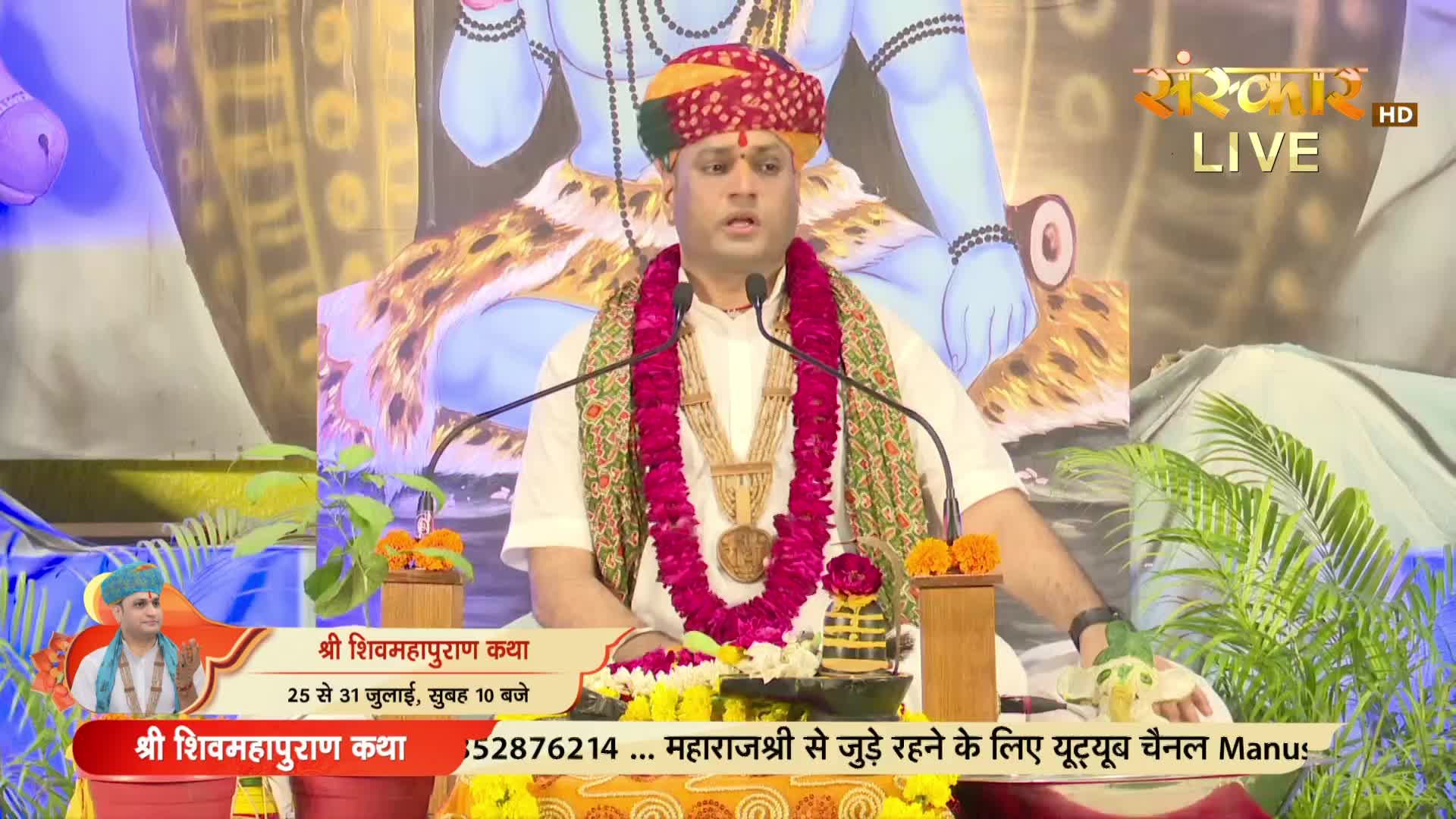 A man in a turban and garland speaks earnestly into two microphones, seated before a backdrop with an image of a deity and the Sanskar TV logo. The broadcast is promoting a religious event in India.
